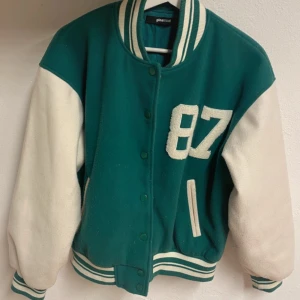 Grön och vit bomberjacka Gina Tricot XS - Snygg bomberjacka i grönt och vitt från Gina Tricot, storlek XS. Jackan har klassisk college-look med broderad 87 på bröstet, ribbade muddar och krage med ränder. Fodrad insida och tryckknappar framtill. Perfekt för en sportig och trendig stil.