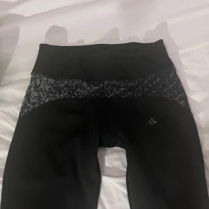 Lågmidjade Svarta Adidas leggings  - Svarta leggings från Adidas med låg midja och snyggt svartvitt mönster över höften. Tillverkade i stretchigt funktionsmaterial, perfekta för träning. Adidas-logga på benet och bekväm passform som sitter tajt längs benen.