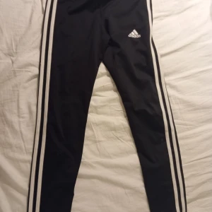 Svarta Adidas träningsbyxor med ränder - Svarta träningsbyxor från Adidasmed klassiska vita ränder längs sidorna och Adidas-logga på låret. Byxorna är tillverkade i ett lätt och stretchigt syntetmaterial (AEROREADY) som andas. Perfekta för sport eller chill. Tight passform. 