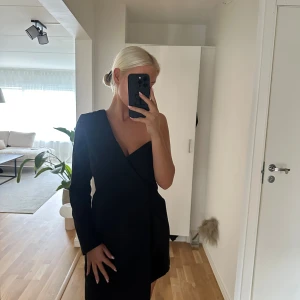 Svart oneshoulder kortklänning från Missguided - Säljer en svart kortklänning från Missguided med unik oneshoulder-design och en lång ärm. Klänningen har en snygg omlott-look framtill och två dekorativa knappar. Materialet är mjukt och känns som polyester. Perfekt för dig som vill sticka ut med en cool och modern stil.