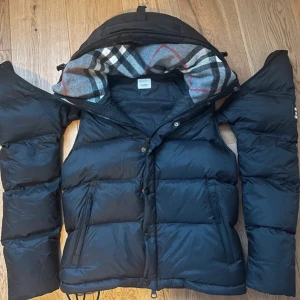 Svart pufferjacka från Burberry - Snygg svart pufferjacka från Burberry med avtagbara ärmar och klassiskt rutigt foder i huvan. Jackan har dragkedja, knappar och två fickor framtill. Perfekt för kalla dagar och riktigt stilren med tydlig Burberry-logga på ärmen.