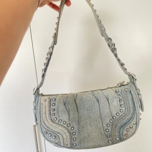 Zara Ljusblå jeans axelväska med nitar - Säljer en ljusblå axelväska i jeansmaterial med coola silverfärgade nitar och ringdetaljer på axelbandet. Väskan är från Zara och den sludsåld. Nypris 599kr. 