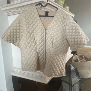 Beige quiltad jacka med dragkedja - Quiltad beige jacka från H&M med korta ärmar och oversized passform. Jackan har ett rutigt mönster, dragkedja framtill och är tillverkad i glansig polyester. Perfekt för lager-på-lager och ger en cool vibe till din outfit.