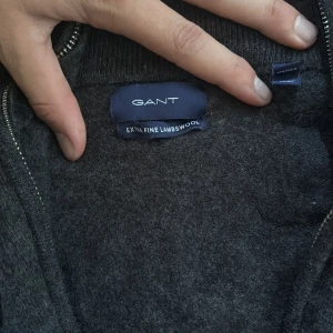 Mörkgrå ullkofta från GANT med dragkedja - Säljer en mörkgrå kofta från GANT i extra fin lammull. Koftan har hel dragkedja framtill, ribbade muddar och broderad GANT-logga på bröstet. Perfekt för lager-på-lager under kyliga dagar.