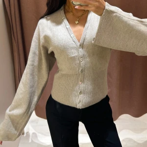 Beige kofta med vida ärmar - Superfin beige kofta med v-ringning och bred ribbad midja. Koftan har vida, långa ärmar och knäpps med pärlemorknappar framtill. Perfekt för lager-på-lager och ger en chill men ändå stilren vibe.