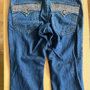 Studded True Religion Ricky T - Säljer ett par blå jeans från True Religion med raka ben och ikoniska bakfickor dekorerade med silvriga nitar. Jeansen har kontrastsömmar och klassisk läderpatch bak. Perfekta för dig som vill sticka ut med detaljer och cool stil.