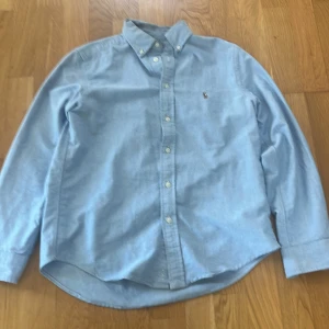 Ljusblå skjorta från Polo Ralph Lauren - Klassisk ljusblå skjorta från Polo Ralph Lauren med broderad logga på bröstet. Skjortan har button-down krage, långa ärmar och knäppning framtill. Tillverkad i mjuk bomull och passar perfekt för en clean och stilren look.