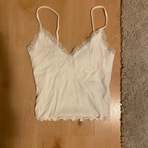 Vit ribbad linnetopp med spets - Söt vit linnetopp med tunna axelband och v-ringning. Toppen är ribbad och har fina spetsdetaljer längs urringningen och upptill. Nederkanten är vågig för en extra touch. Perfekt för sommarens alla outfits!