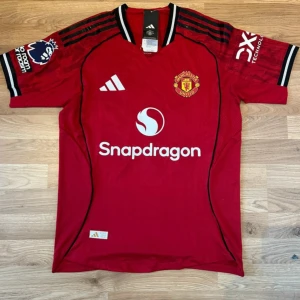 Manchester United matchtröja Cunha 10 - Röd Manchester United fotbollströja från Adidas med tryck 'Cunha 10' på ryggen. Tröjan har korta ärmar, svarta och vita detaljer på axlar och ärmslut samt klubbmärke och sponsorer på bröstet. Tillverkad i lätt och ventilerande funktionsmaterial.