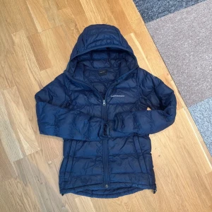 Mörkblå dunjacka från Peak Performance - Snygg mörkblå dunjacka från Peak Performance med huva och dragkedja framtill. Jackan är quiltad och har en sportig look, perfekt för kyliga dagar. Passformen är normal och materialet är lätt och värmande.