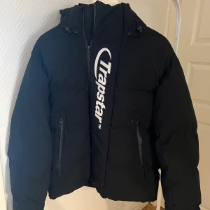 Svart pufferjacka Trapstar S - Svart pufferjacka från Trapstar med stor vit logga på framsidan och svart logga på ryggen. Jackan har huva med snörning, två fickor med dragkedja och en clean, oversized look. Perfekt för dig som vill ha streetstyle-vibe och synas i mängden.