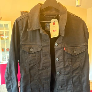 Svart jeansjacka från Levi's - Svart jeansjacka från Levi's med klassisk krage, raka axlar och knappar framtill. Jackan har två bröstfickor med lock och knapp, samt den ikoniska röda Levi's-taggen. Snygg och enkel att matcha med allt.