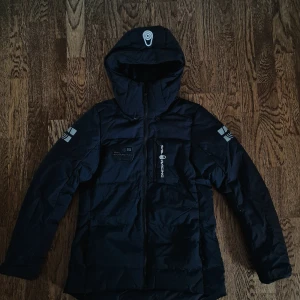 Svart dunjacka från Sail Racing (Dam) - Säljer en svart dunjacka från Sail Racing i storlek S. Jackan har en stilren design med dragkedja och huva. Perfekt för kalla vinterdagar med sitt Allied Feather Down-foder. Perfekt för vintern och hösten. Den är inte använd mer ett fåtal gånger. Orignalpriset ligger på 2499:- men jag säljer för 1500:-