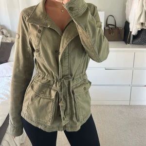 Olivgrön jacka med snörning - Säljer en olivgrön parkajacka från hollister med snygg snörning i midjan och stora fickor framtill. Jackan har långa ärmar, knappar och dragkedja samt en cool patch på ena ärmen. Perfekt för dig som gillar streetstyle och vill ha en chill look.