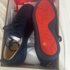 Mörkblå sneakers från Louboutin - Säljer ett par mörkblå sneakers från Christian Louboutin i mocka med den ikoniska röda sulan. Skorna har rund tå, platt sula och snörning. Insidan är beige och de levereras i originalkartong med tillhörande papper. Perfekt för dig som vill sticka ut med lyxiga detaljer.