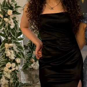 Svart långklänning med slits - Säljer en elegant svart långklänning i satinliknande material. Klänningen är axelbandslös med figurnära passform och har en hög slits på ena sidan som ger en snygg och modern look. Perfekt för dig som vill sticka ut på bal eller fest.