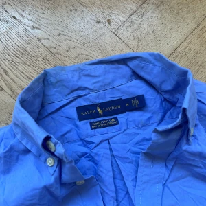 Blå skjorta från Ralph Lauren M - Snygg blå skjorta från Ralph Lauren i slim fit med klassisk krage och knappar framtill. Skjortan är tillverkad i 100% bomull med stretch och har den ikoniska broderade loggan på bröstet. Perfekt för dig som gillar stilrena och tidlösa plagg.