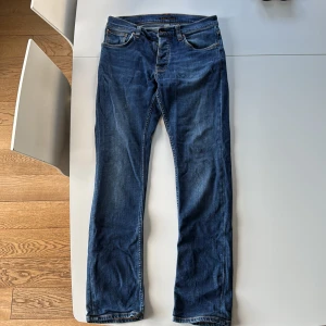 Blå jeans Grim Tim från Nudie Jeans, stl 31/32 - Snygga blå jeans från Nudie Jeans med klassisk femficksdesign och orange sömmar. Innerbensmått 79cm, över midja 40cm. Färg indigo myth. Jeansen har raka ben och normal passform, samt ikonisk broderad detalj på bakfickorna. Perfekta för dig som gillar stilrena och tidlösa jeans.