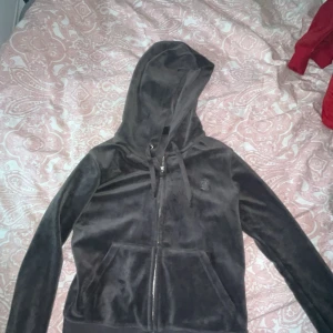 Mörk grå velour hoodie Juicy Couture - Mysig mörkgrått hoodie från Juicy Couture i mjuk velour med dragkedja framtill och huva med snörning. Klassisk logga broderad på bröstet och två stora fickor framtill. Perfekt för chill dagar eller när du vill vara extra comfy.