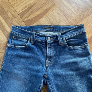 Blå skinny jeans från Nudie Jeans - Snygga blå jeans från Nudie Jeans med klassisk femficksdesign och orangea kontrastsömmar. Modellen är skinny fit med smala ben och normal midja. Jeansen har Nudie Jeans signaturbrodyr på bakfickorna och är tillverkade i mjukt denimtyg.