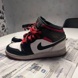 Nike Air Jordan 1 Mid svart/vit/röd - Nike Air Jordan 1 Mid sneakers i svart, vit och röd med klassisk Jordan-logga på sidan.  Perfekt för dig som gillar streetwear och ikoniska sneakers. Använd hyfsat mycket men annars väldigt fina 