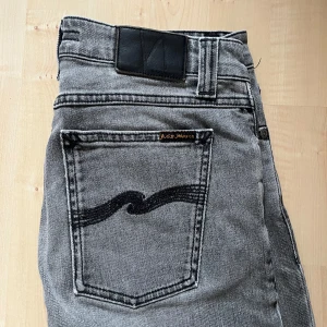 Grå jeans från Nudie Jeans - Säljer ett par grå jeans från Nudie Jeans med klassisk femficksdesign och svarta broderade detaljer på bakfickan. Jeansen har en smal passform och är tillverkade i mjukt denimtyg. Perfekta för dig som gillar en stilren och modern look.