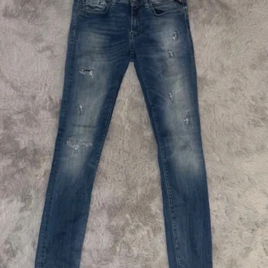 Replay blå jeans med slitningar - Tvärfeta  blå jeans från Replay i anbass modellen med coola slitningar och tvättade detaljer. Klassisk femficksmodell med knapp och dragkedja.