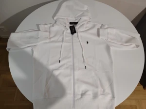 Vit hoodie från Polo Ralph Lauren - Snygg vit hoodie från Polo Ralph Lauren med dragkedja, huva med dragsko och klassisk broderad logga på bröstet. Stora fickor framtill och ribbade muddar. Perfekt för en clean och stilren look. Finns i olika storlekar, S,M,L