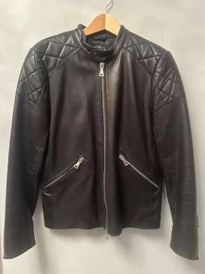 Svart skinnjacka Jofama x King - Snygg svart skinnjacka från Jofama x King med quiltade detaljer på axlar och ärmar. Jackan har ståkrage med knapp, två snedställda fickor med dragkedja och dragkedja även vid ärmslut. Perfekt för dig som gillar stilren och edgy look.