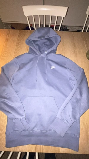 Nike lila hoodie med logga - Snygg lila hoodie från Nike med klassisk vit logga på bröstet. Hoodien har en stor känguruficka framtill, ribbade muddar och justerbar dragsko i huvan. Mjuk insida i bomullsblandning som är skön mot huden. Perfekt för chill eller streetwear.
