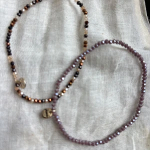 Två pärlarmband från Pilgrim  - Säljer två snygga pärlarmband från Pilgrim. Ett armband har små lila pärlor och ett har pärlor i svart, guld och beige med ett coolt mönster. Båda har en liten guldbricka med logga. Perfekt att kombinera för en personlig stil.