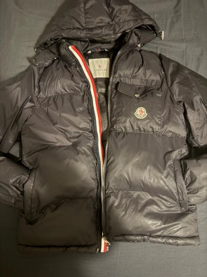 Svart Moncler Montbeliard S - Svart pufferjacka från Moncler med huva och klassisk logga på bröstet. Jackan har dragkedja med röd, vit och blå kant, samt en ficka med knapp framtill. Fylld med vitt gåsdun för extra värme. Perfekt för kalla dagar och snygg streetstil.