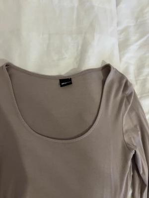 Beige långärmad topp Gina Tricot - En stilren beige långärmad topp från Gina Tricot med rundad, djup urringning. Tunn och mjuk i materialet, perfekt att styla med jeans eller kjol. Enkel design som funkar till många looks.