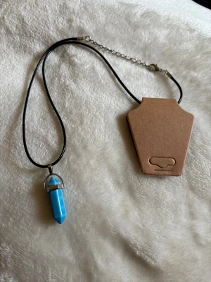 Halsband med turkos kristallhänge - Snyggt halsband med svart läderimitation och ett turkost kristallformat hänge i metallinfattning. Justerbar kedja i silverfärg gör att längden kan anpassas. Perfekt accessoar för att ge din outfit en boho-touch.