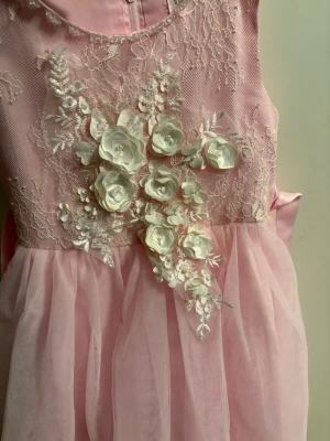 Rosa prinsessklänning med spets och blommor - Superfin rosa prinsessklänning med ärmlös design, spets upptill och stora vita 3D-blommor med pärlor framtill. Kjolen är fluffig i flera lager och baktill finns ett brett band som knyts till rosett. Perfekt för ditt barn som vill sticka ut med romantiska detaljer och mjuk färg.