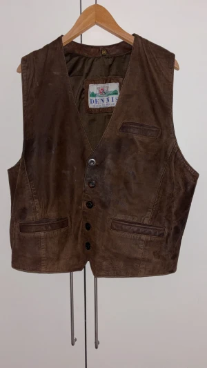 Brun skinnväst från Dennis Päls & Skinn - Snygg brun skinnväst från Dennis Päls & Skinn AB med klassisk v-ringning och knäppning framtill. Västen har flera fickor och är tillverkad i äkta skinn för en tidlös look. Perfekt för dig som vill addera lite edge till din outfit.