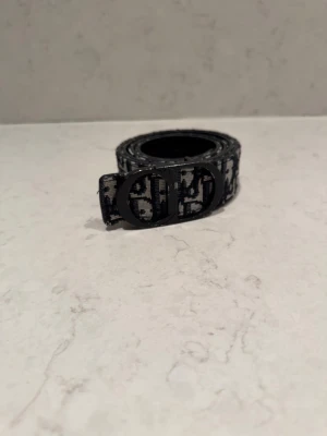 Svart och vit bälte med monogram - Snyggt svart och vitt bälte med ikoniskt monogrammönster och rundad metallspänne med CD-logga. Bältet har en stilren design och är tillverkat i skinn med textil på ovansidan. Perfekt accessoar för att lyfta din outfit.