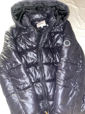 Svart pufferjacka från ONLY med huva - Säljer en svart pufferjacka från ONLY med huva och dragkedja framtill. Jackan har quiltad design, två fickor med tryckknappar och en badge på ärmen. Perfekt för kalla dagar och riktigt skön att bära.