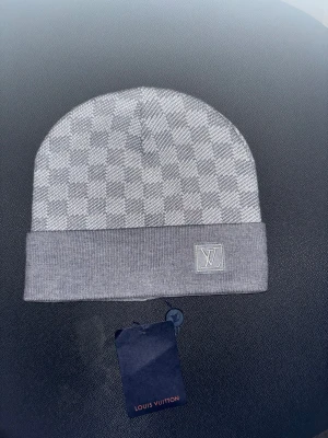 Grå rutig mössa - En elegant och varm mössa i mjuk kvalitet, designad i ett grått rutmönster som ger en exklusiv och modern look. Den ribbstickade uppvikskanten ger både bättre passform och extra komfort. Perfekt för kalla dagar och passar lika bra till vardagsstil som till mer uppklädda outfits. Ett stilrent val för dig som vill kombinera värme med minimalistisk design.