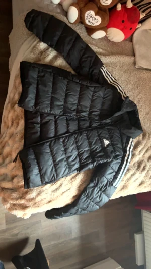 Svart pufferjacka från Adidas - Säljer en svart pufferjacka från Adidas med klassiska vita ränder längs ärmarna och Adidas-logga på bröstet. Jackan har dragkedja framtill, hög krage och quiltad design. Perfekt för kalla dagar och riktigt snygg streetstil.