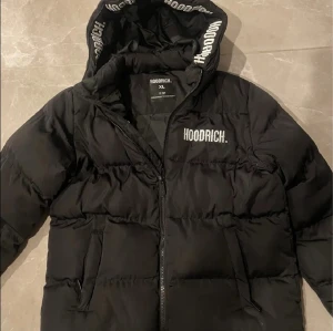 Svart pufferjacka från Hoodrich - Svart pufferjacka från Hoodrich i storlek XS (13-16 år), med huva och logga tryckt på bröstet och insidan av huvan. Jackan har dragkedja framtill och två sidofickor. Perfekt för kalla dagar med sin tjocka och vadderade design. Använts i 1 månad så den är i mycket bra skick!