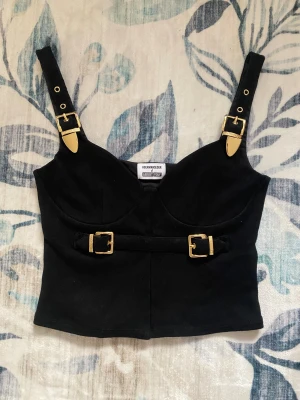 About You topp - Svart, korsettliknande topp från About You med guldfärgade bucklor i storlek L. Originalpris: ca 400 kr. Tjockt tyg, ganska ”tung” pga bucklorna, därav fraktpriset. Endast testad och därav i prima skick. 