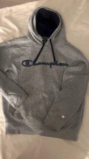 Grå Champion hoodie  - Säljer en grå unisex Champion-hoodie (ändå lite oversized än xs )i riktigt bra skick. Inga fläckar och inget slitage. kvalitén är fortfarande topp trots användning. Materialet är mjukt och skönt, och hoodien har en praktisk magficka framtill.  Stilren design med mörkblå Champion text på bröstet. Passar lika bra till gymmet som till vardags, och är både bekväm och hållbar.