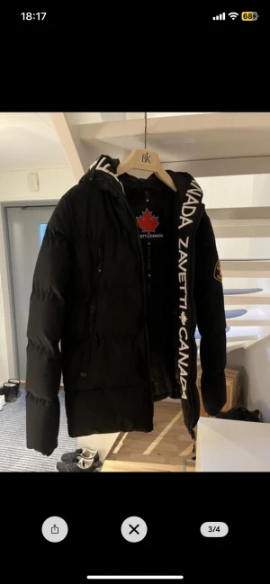 Svart pufferjacka från Zavetti Canada - Säljer en svart pufferjacka från Zavetti Canada med huva och tydlig logga på ärmen. Jackan har dragkedja framtill och textdetaljer längs hela jackan och insidan av huvan. Perfekt för kalla dagar och har en schysst, modern look. Hör av er vid frågor!