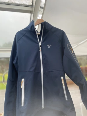 Marinblå vindjacka från Tenson L - Snygg marinblå vindjacka från Tenson med vit dragkedja och coola detaljer på ärmen. Jackan har hög krage, tre zip-fickor med orangea dragare och sportig look. Perfekt för dig som gillar outdoor och vill ha en stilren jacka med funktion.