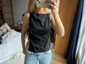 Ärmlös topp med knyt - Svart ärmlös topp med öppen sida som knyts med tunna band. Toppen har en enkel, stilren design 