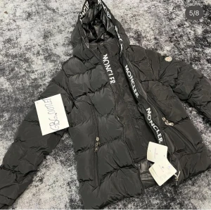 Svart Moncler pufferjacka med huva - Svart pufferjacka från Moncler med stor huva och logotypband. Jackan har två framfickor med dragkedja, ribbade muddar och klassisk Moncler-märke på ärmen. Materialet är glansig polyester och dunfyllning, perfekt för kalla dagar. Snygg och modern look.