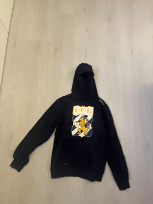 Svart IFK Göteborg hoodie från Craft - Svart hoodie från Craft med stort IFK Göteborg-lejon på bröstet. Tröjan har huva, lång ärm och ribbade muddar. Materialet är mjukt och passar perfekt för dig som gillar sport och vill visa ditt lagstöd. Snygg och enkel design med Craft-logga på axeln.