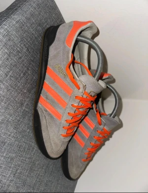 Adidas Jeans sneakers grå och orange - Säljer ett par Adidas Jeans sneakers i mocka med grå bas och orangea detaljer. Skorna har klassiska tre ränder på sidan, orange snörning och svart sula. Guldtexten 'Jeans' syns på sidan. Perfekta för dig som gillar retrostil och färgklickar.