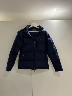 Mörkblå pufferjacka från Polo Ralph Lauren - Snygg mörkblå pufferjacka från Polo Ralph Lauren med röd broderad logga på bröstet. En av dem mest eftertraktade jackorna på marknaden! Perfekt nu till vinter, både snygg och håller dig varm! Bara skriva till mig om ni har några frågor eller funderingar!